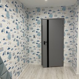 Wojciech Wałek - Pokój dziecięcy po remoncie: szara, nowoczesna drzwi z czarnym akcentem na tle ściany pokrytej tapetą w dziecięce wzory z autami i budynkami, jasna podłoga imitująca drewno.