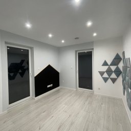 Wojciech Wałek - Pusty, jasny pokój po remoncie z panelami akustycznymi na ścianach: czarny panel w kształcie domku i kompozycja trójkątów w odcieniach szarości, jasna podłoga imitująca drewno, białe ściany i sufit...