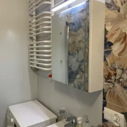 Nowoczesna łazienka z białą pralką, umywalką z baterią i dozownikiem mydła, grzejnikiem drabinkowym, szafką z lustrem podświetlaną LED oraz tapetą w kwiaty.