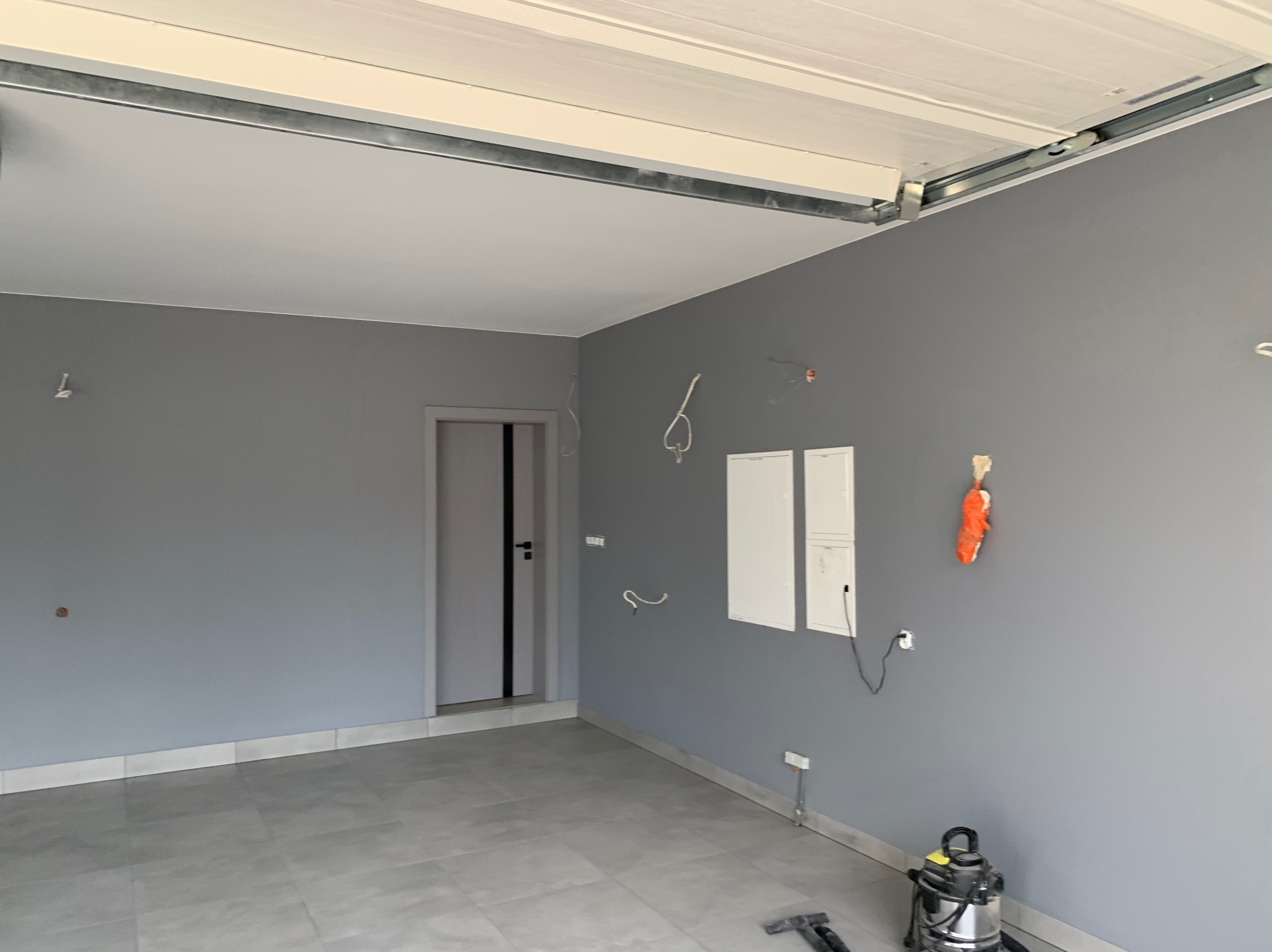 Szare wnętrze pokoju po malowaniu ścian i ułożeniu szarych płytek podłogowych, widoczne niedokończone instalacje elektryczne i odkurzacz przemysłowy.