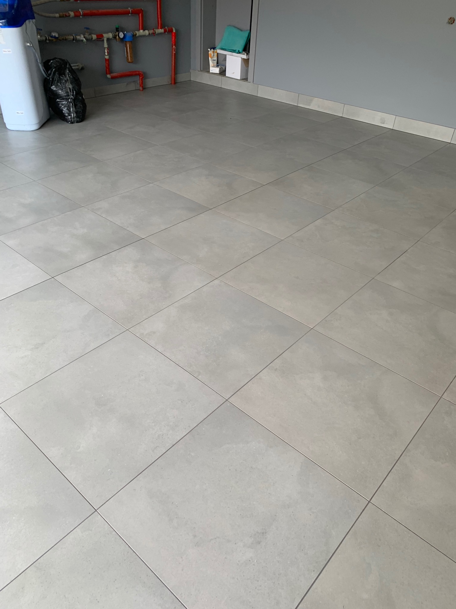 Jasnoszara podłoga wyłożona kwadratowymi płytkami, widoczne rury instalacyjne pomalowane na czerwono oraz biały zmiękczacz wody i worek na śmieci.