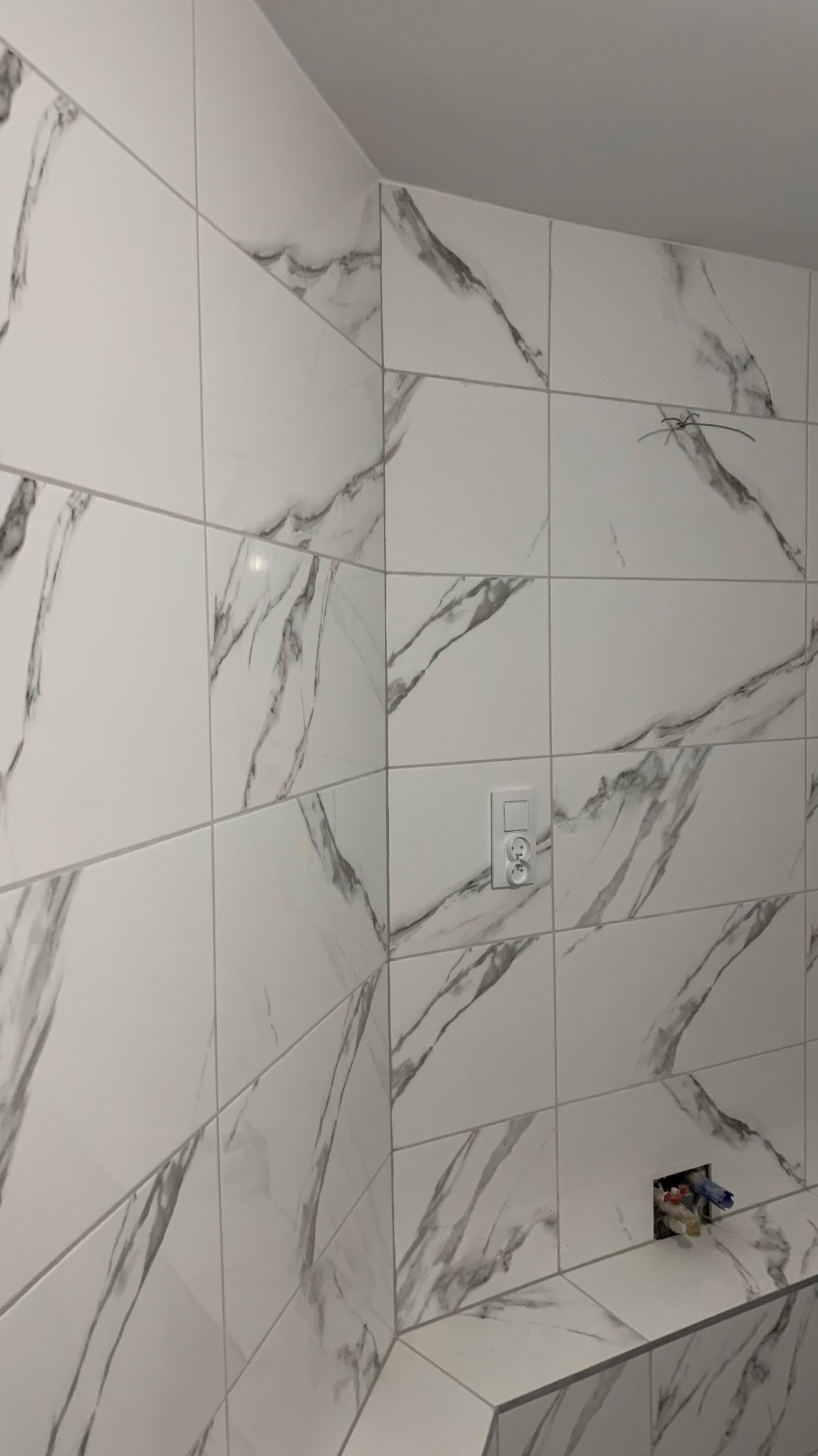 Ściana wyłożona białymi płytkami imitującymi marmur, widoczne gniazdko elektryczne i wystające rurki instalacji wodnej.