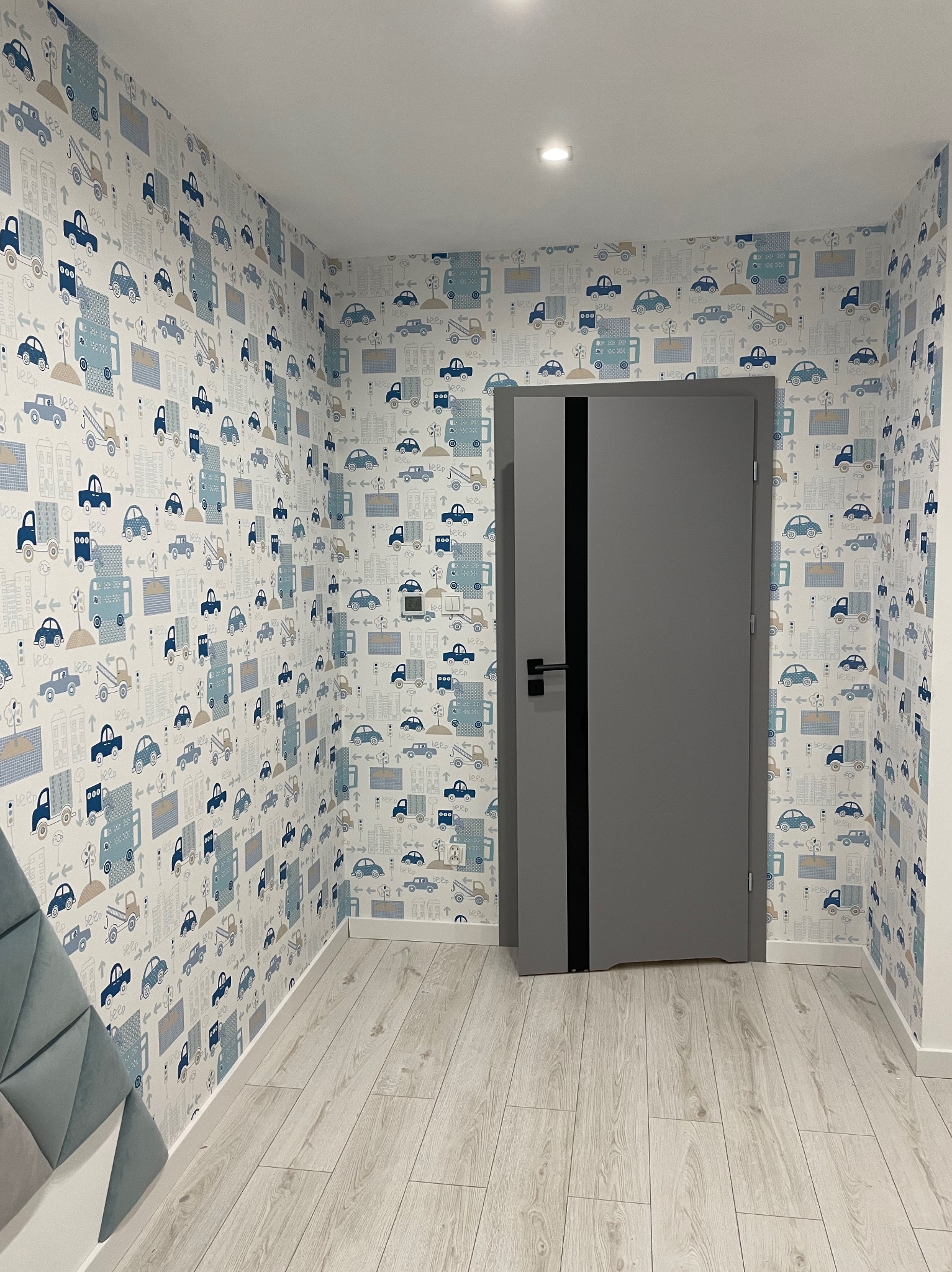 Pokój dziecięcy po remoncie: szara, nowoczesna drzwi z czarnym akcentem na tle ściany pokrytej tapetą w dziecięce wzory z autami i budynkami, jasna podłoga imitująca drewno.