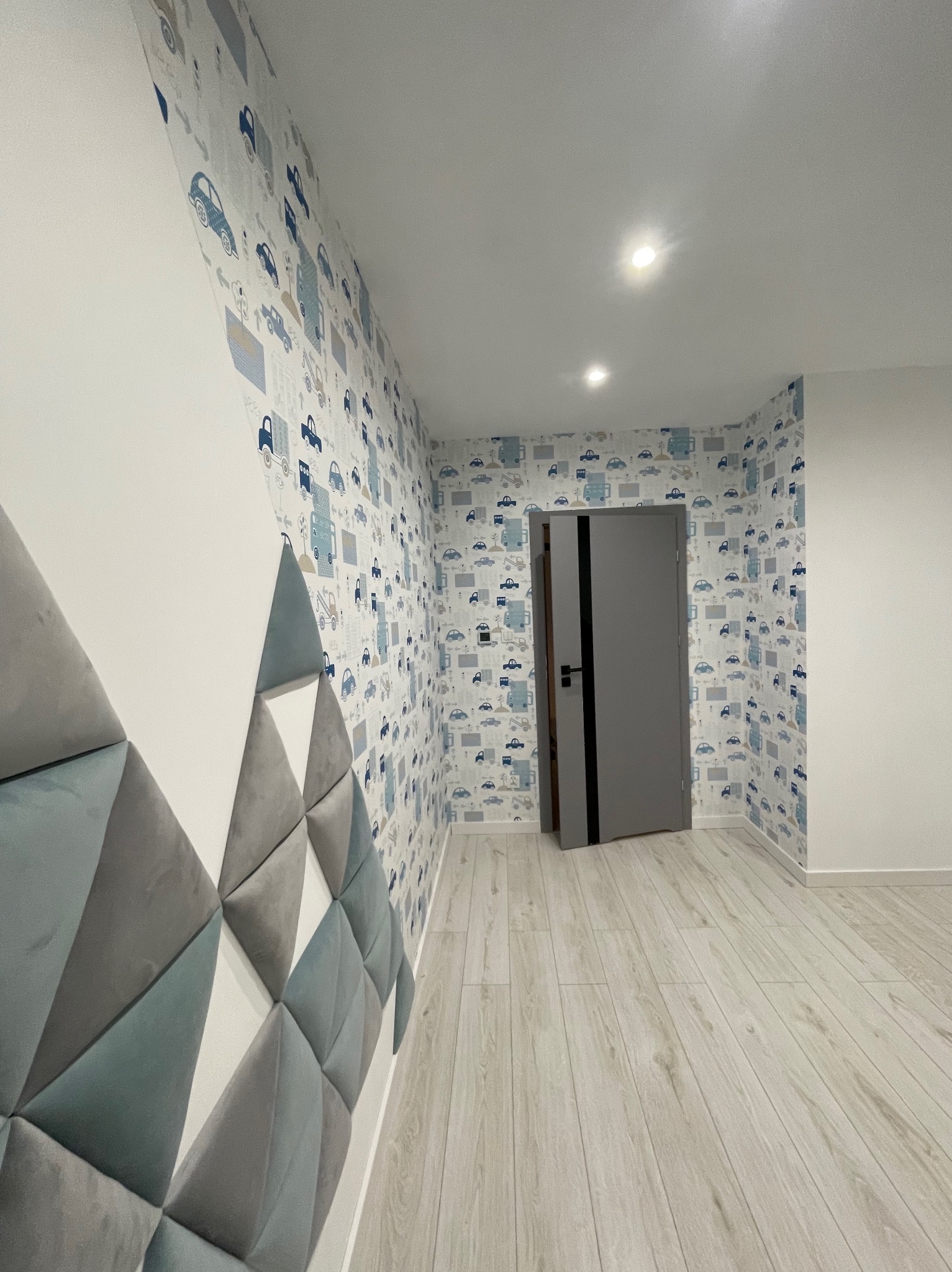 Pokój dziecięcy po remoncie: ściana z tapetą w samochodziki, miękkie panele ścienne w kształcie trójkątów w odcieniach szarości i błękitu, szare drzwi z czarnym paskiem, jasna podłoga imitująca...
