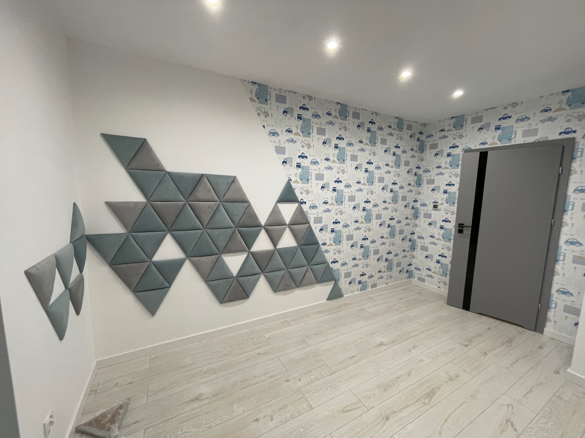 Pokój po remoncie z tapetą w dziecięce motywy samochodów, ścianą z trójkątnych paneli akustycznych w odcieniach szarości i bieli oraz szarą ościeżnicą drzwi.