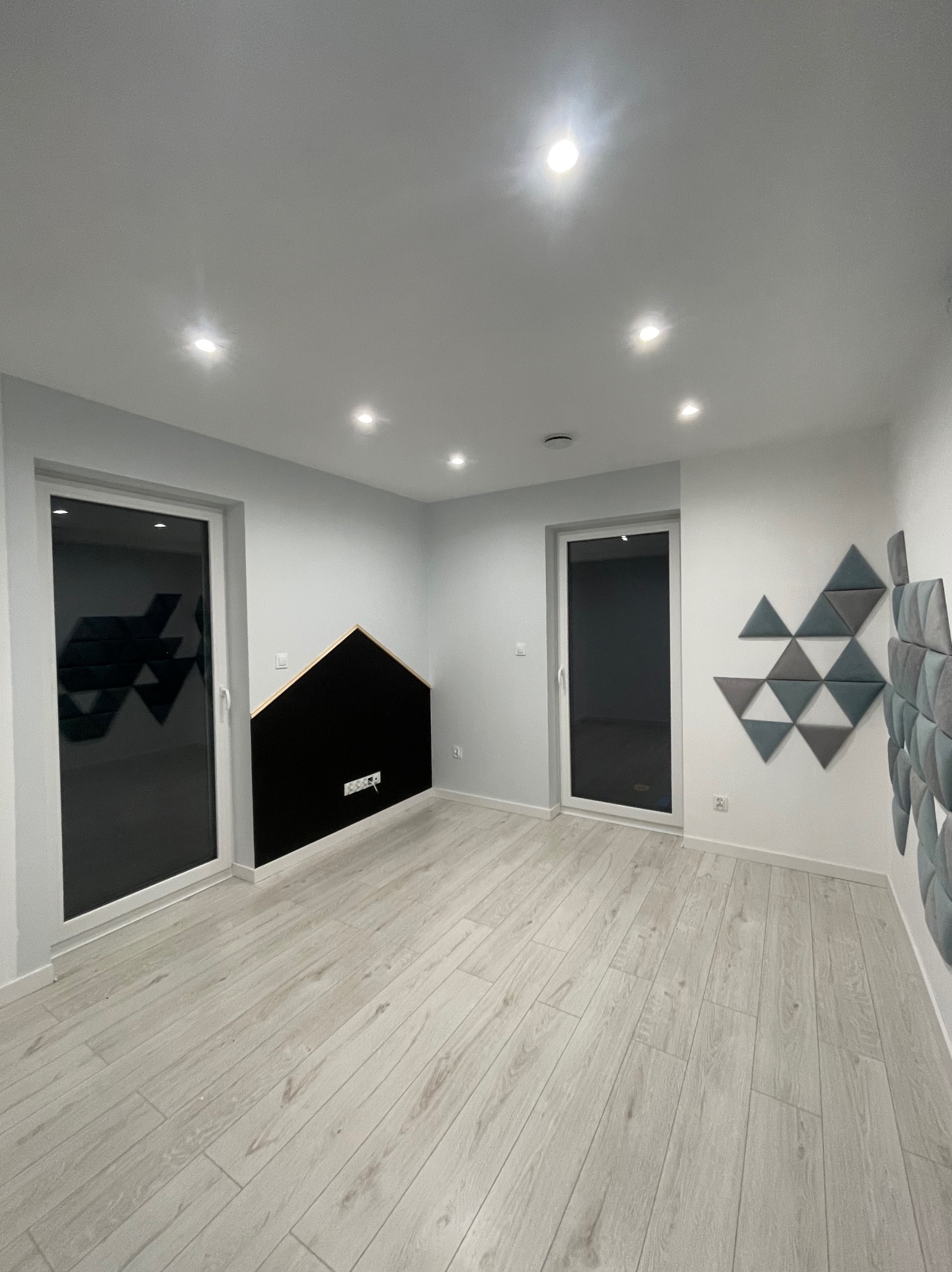 Pusty, jasny pokój po remoncie z panelami akustycznymi na ścianach: czarny panel w kształcie domku i kompozycja trójkątów w odcieniach szarości, jasna podłoga imitująca drewno, białe ściany i sufit...