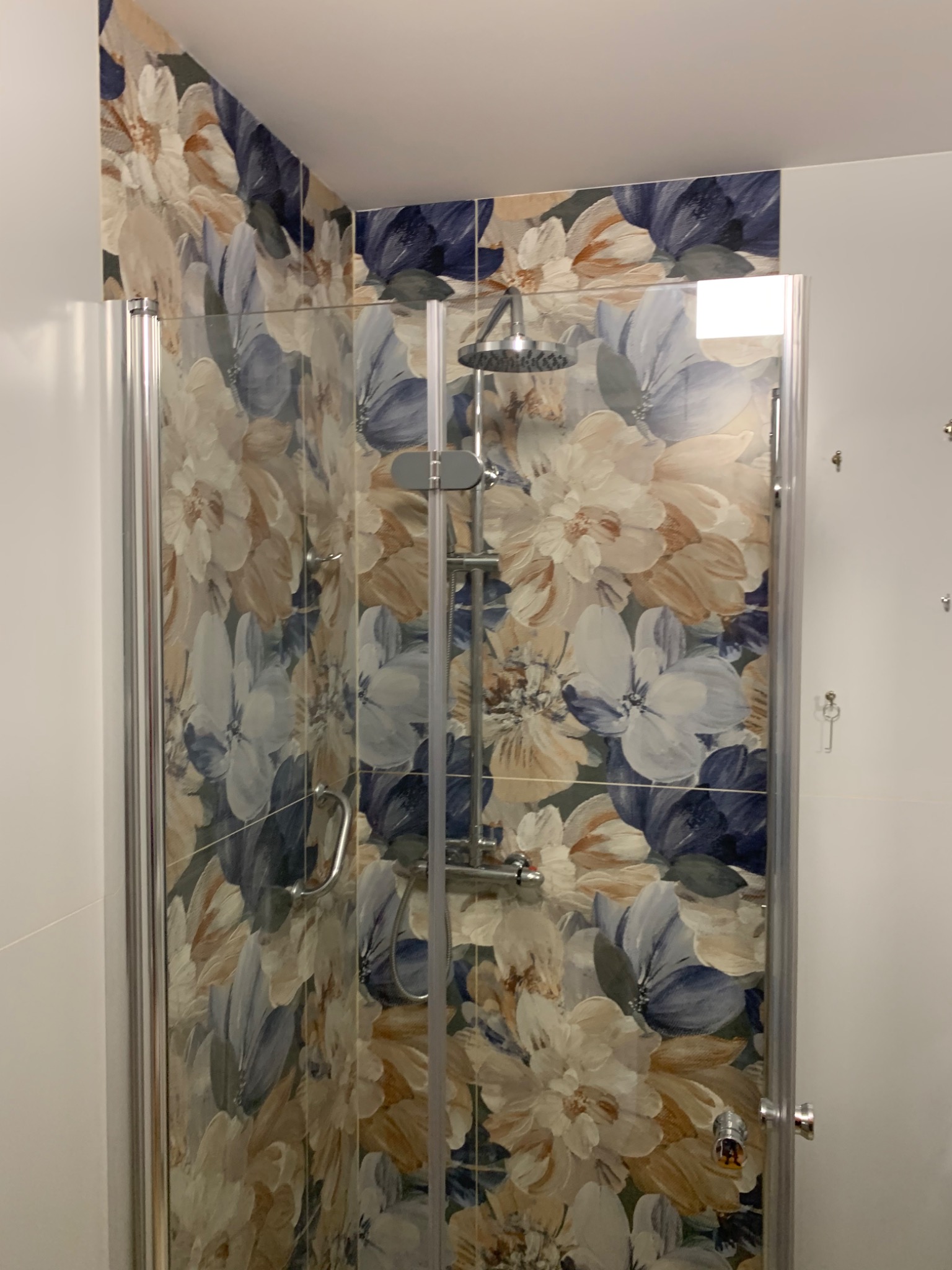 Kabina prysznicowa z motywem kwiatowym na płytkach, chromowana armatura i szklane drzwi, widok z zewnątrz.
