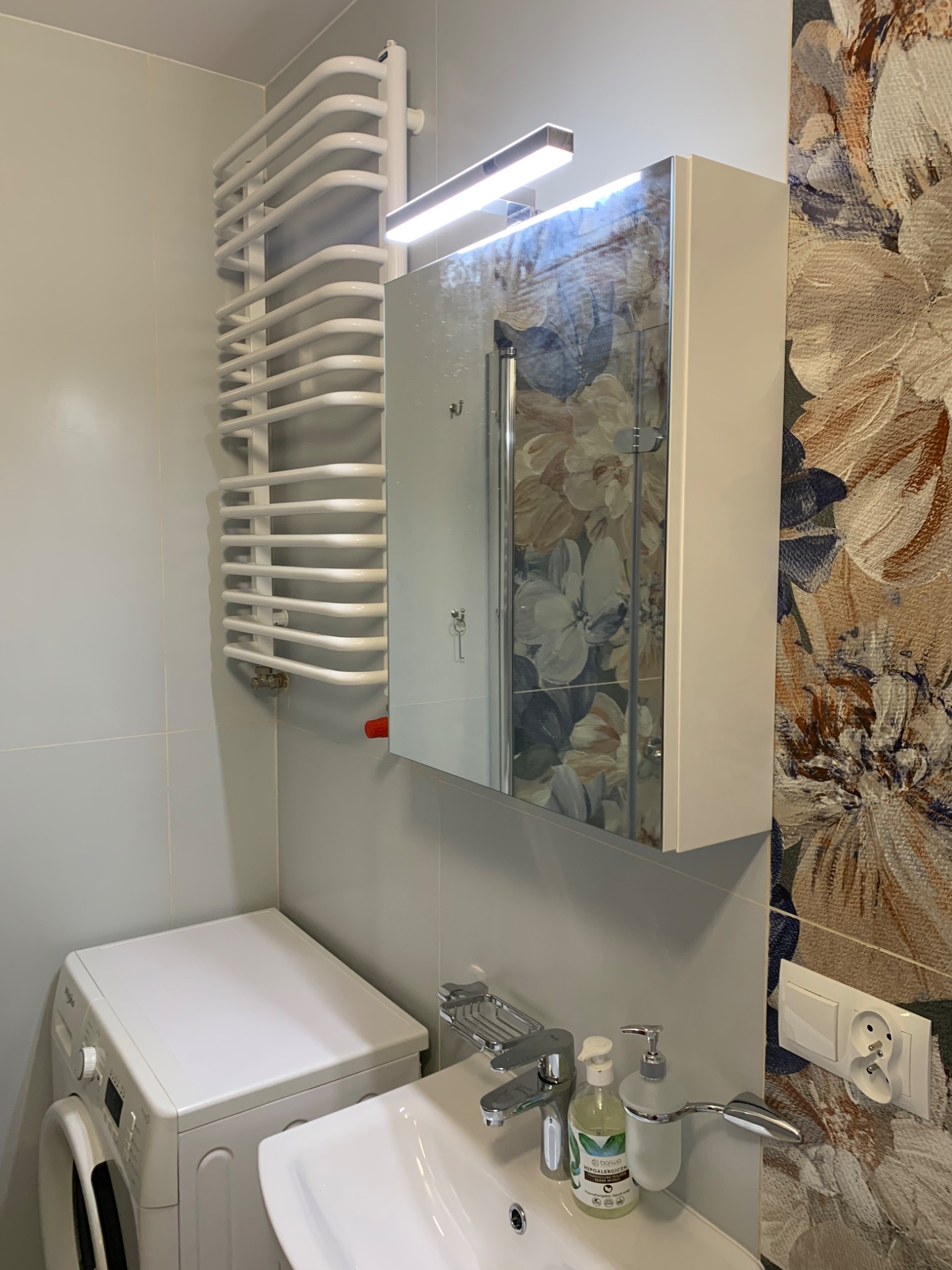 Nowoczesna łazienka z białą pralką, umywalką z baterią i dozownikiem mydła, grzejnikiem drabinkowym, szafką z lustrem podświetlaną LED oraz tapetą w kwiaty.