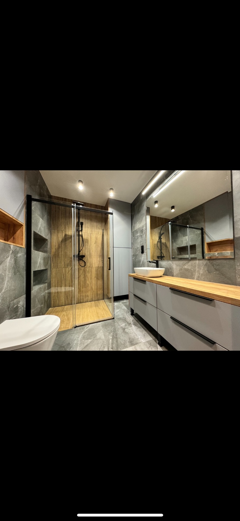 Nowoczesna łazienka z kabiną prysznicową typu walk-in z drewnianym panelem, szarą marmurową płytką, umywalką nablatową na szafce z drewnianym blatem i dużym lustrem z podświetleniem.