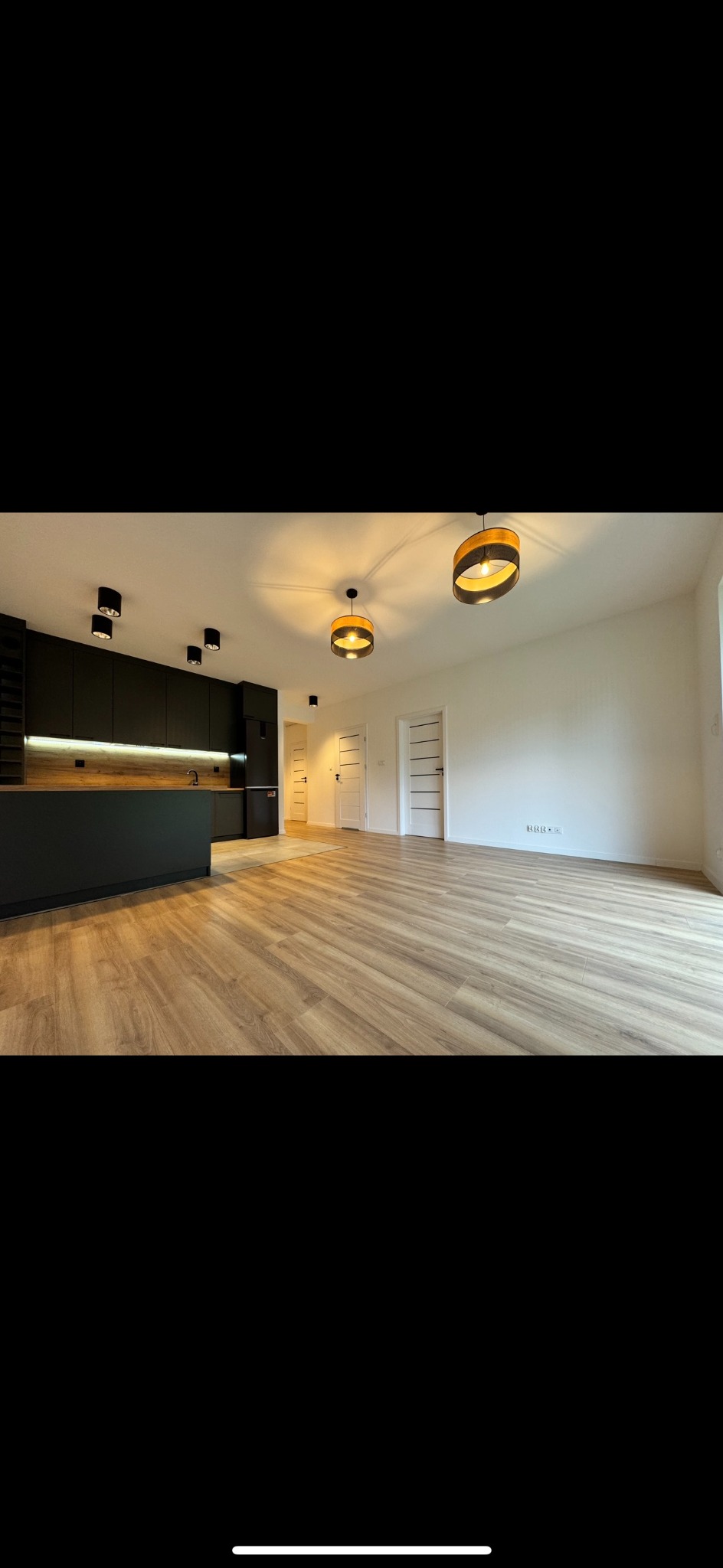 Salon z panelami podłogowymi w odcieniach drewna, czarną zabudową kuchenną z drewnianym blatem i nowoczesnym oświetleniem w postaci dwóch wiszących lamp oraz czarnych reflektorów sufitowych.