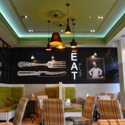 Yurii Fedenko Solutions - Wnętrze pizzerii z zielonymi lożami, krzesłami w kratę, czarną ścianą z rysunkami sztućców i napisem 'EAT PIZZA' oraz portretem kucharza w ozdobnej ramie.