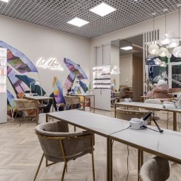 Yurii Fedenko Solutions - Jasne wnętrze salonu kosmetycznego z marmurowymi blatami, złotymi ramami i krzesłami, dekoracją ścienną z motywem tropikalnych liści i neonowym napisem 'be More'.