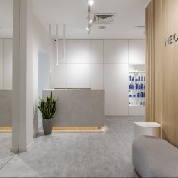 Yurii Fedenko Solutions - Jasne, minimalistyczne wnętrze biura z recepcją, rośliną doniczkową i podświetlanym logo firmy 'NEOLASER' na drewnianej ścianie. W tle widoczna przeszklona witryna z produktami kosmetycznymi.