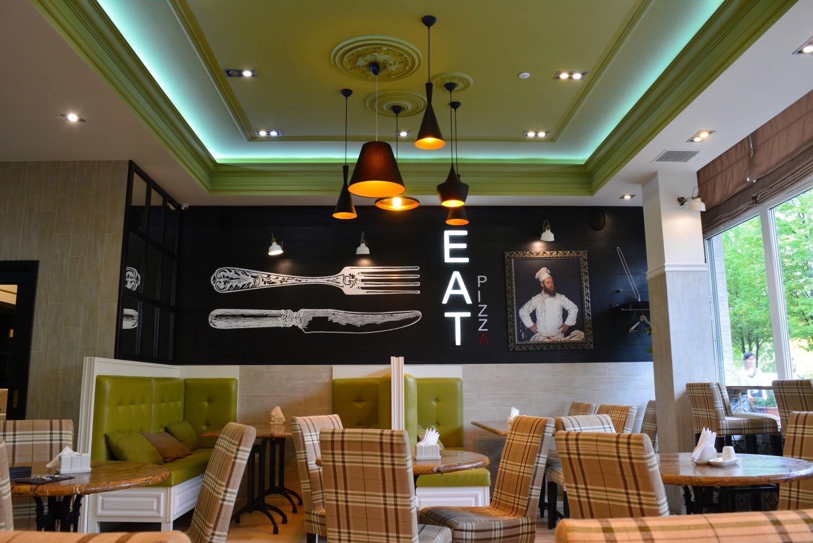 Wnętrze pizzerii z zielonymi lożami, krzesłami w kratę, czarną ścianą z rysunkami sztućców i napisem 'EAT PIZZA' oraz portretem kucharza w ozdobnej ramie.