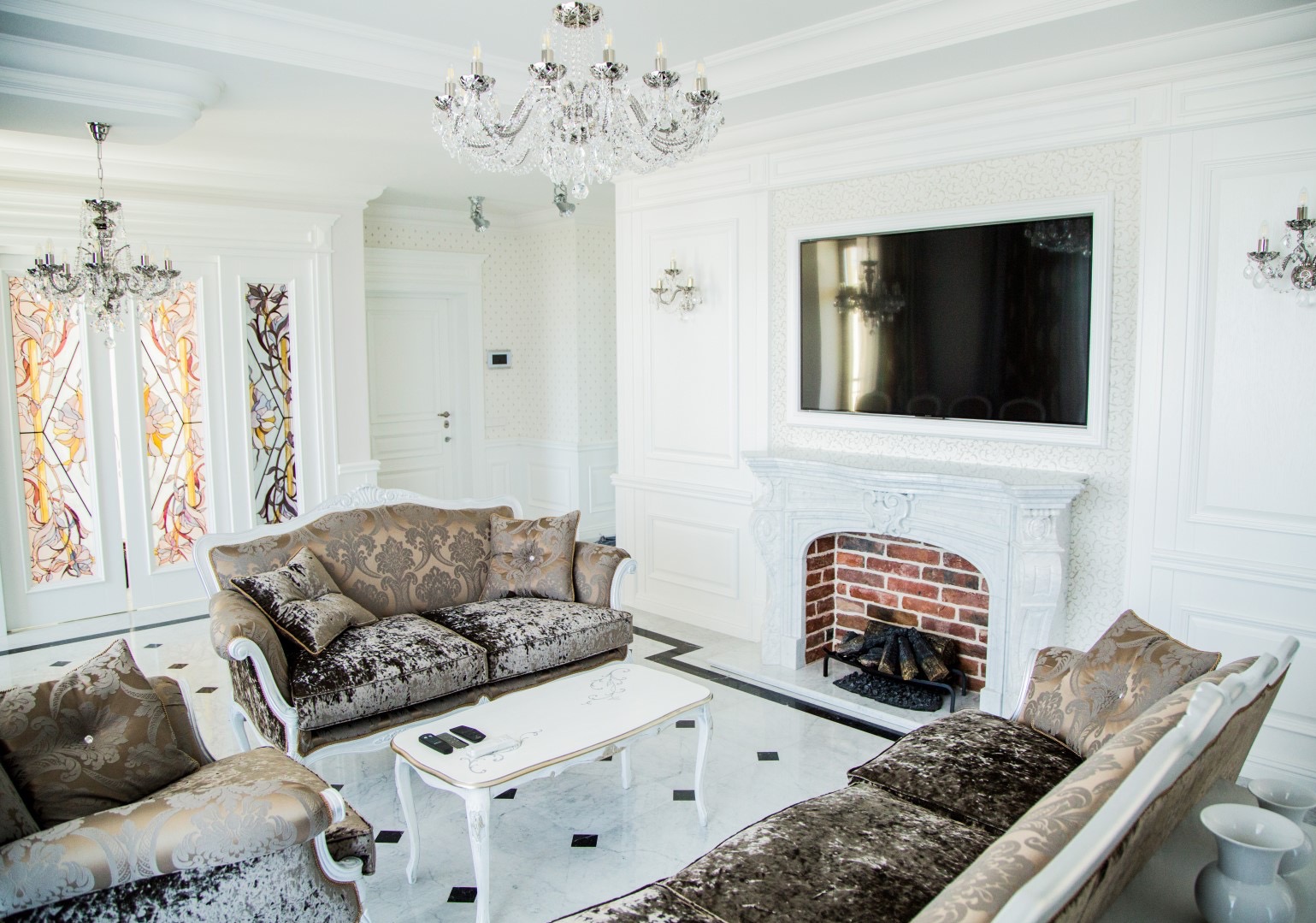 Elegancki salon w stylu glamour z marmurową podłogą, kryształowymi żyrandolami, kominkiem z ceglanym wnętrzem i telewizorem w białej ramie, sofa i fotel z wzorzystą tapicerką.