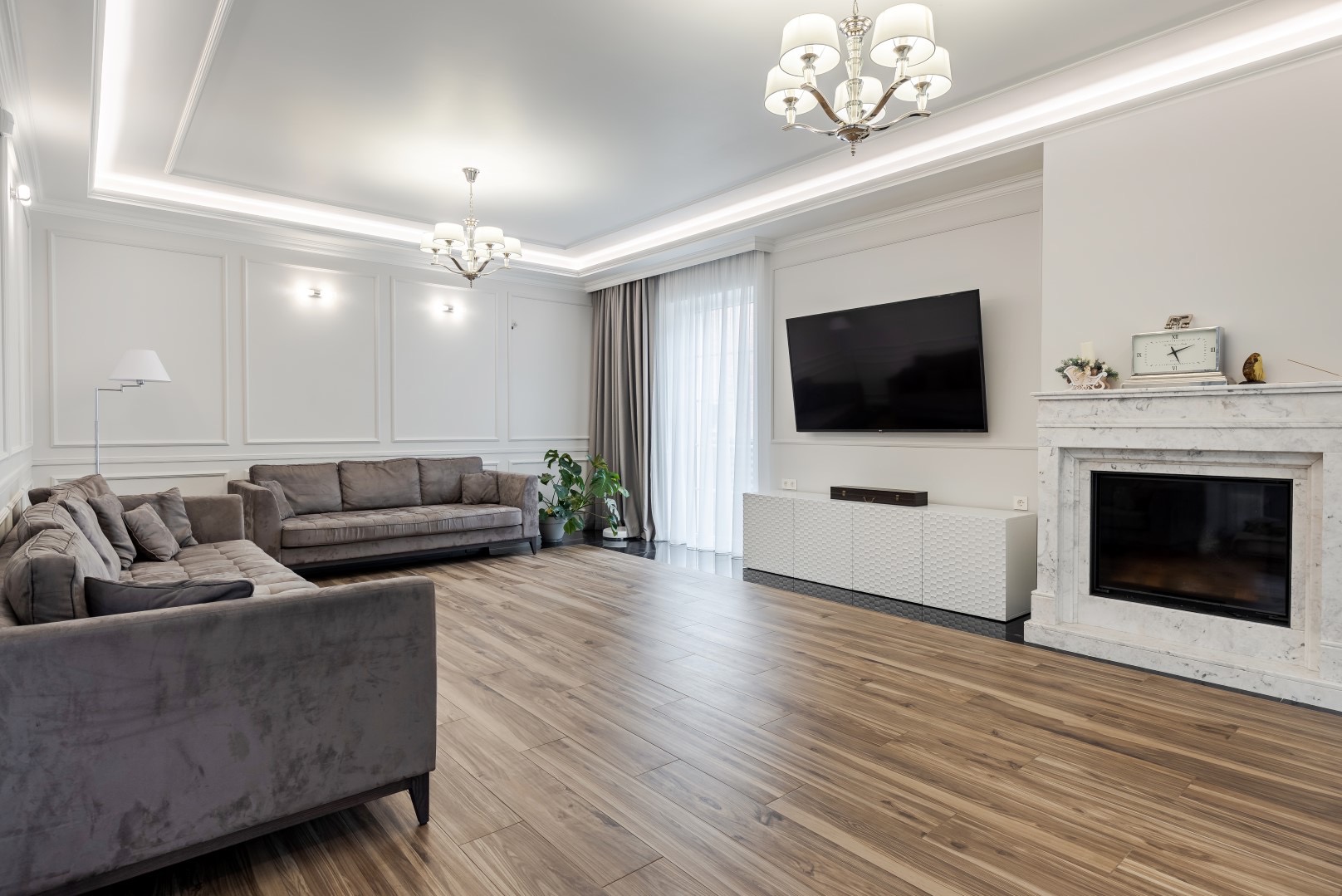 Jasny salon z drewnianą podłogą, szarą sofą narożną i prostą sofą, białymi ścianami z dekoracyjnymi panelami, marmurowym kominkiem z zegarem, telewizorem na ścianie i białym kredensem...