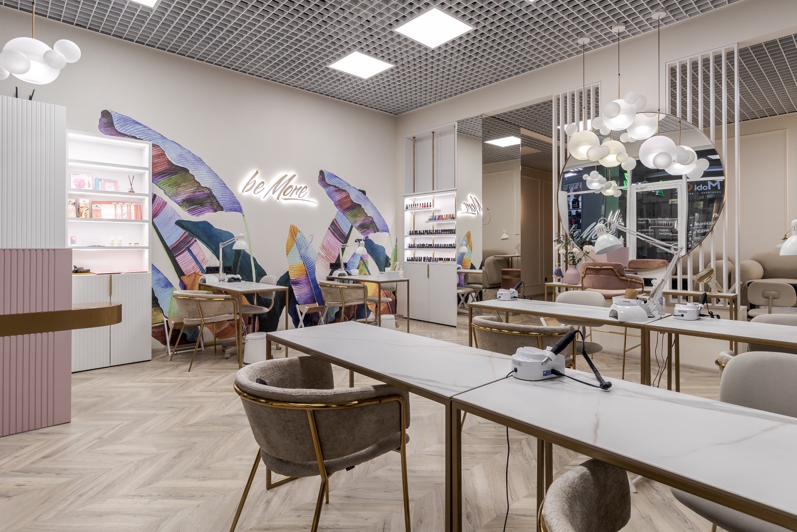Jasne wnętrze salonu kosmetycznego z marmurowymi blatami, złotymi ramami i krzesłami, dekoracją ścienną z motywem tropikalnych liści i neonowym napisem 'be More'.