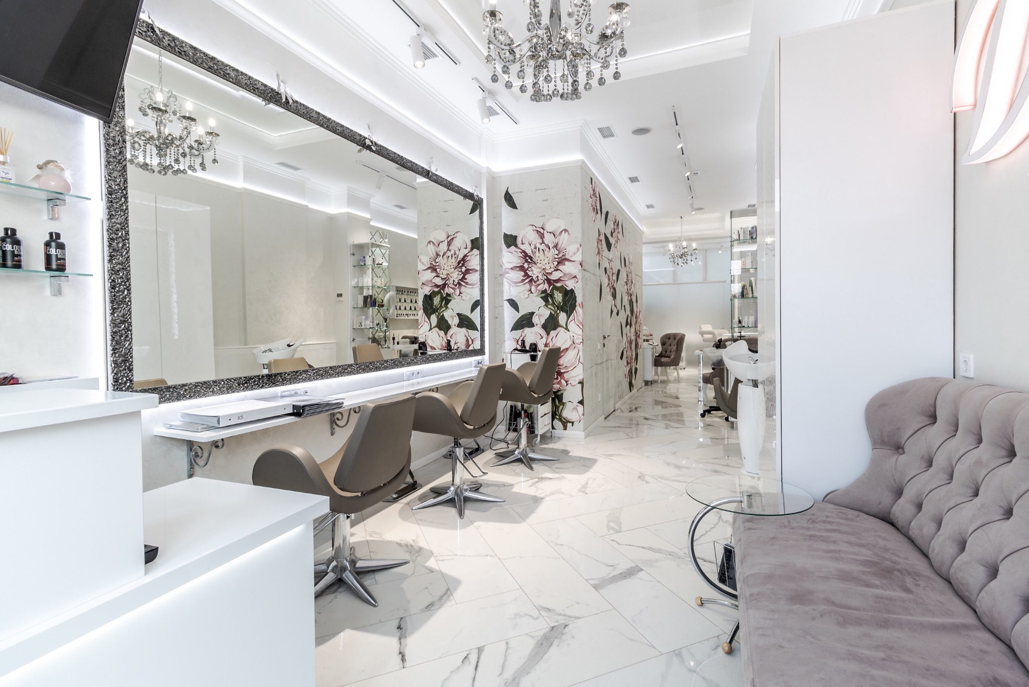 Jasne, przestronne wnętrze salonu kosmetycznego z marmurową podłogą, dużym lustrem w ozdobnej ramie, tapetą w kwiaty i wygodną sofą dla klientów.
