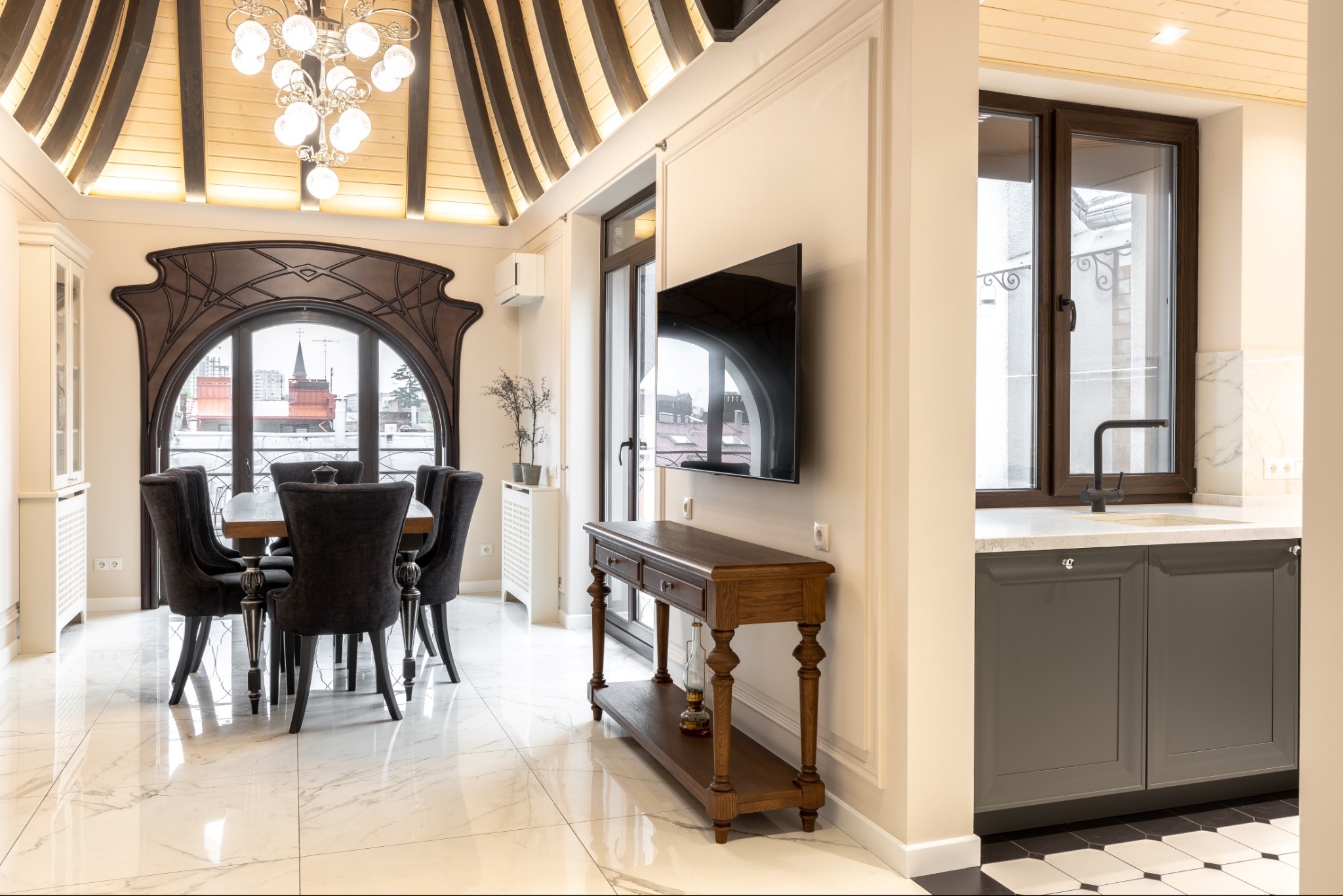 Eleganckie wnętrze mieszkania: marmurowa podłoga, stół jadalny z krzesłami, drewniana konsola pod telewizorem, designerskie okno z widokiem na miasto i fragment kuchni z szarymi szafkami.