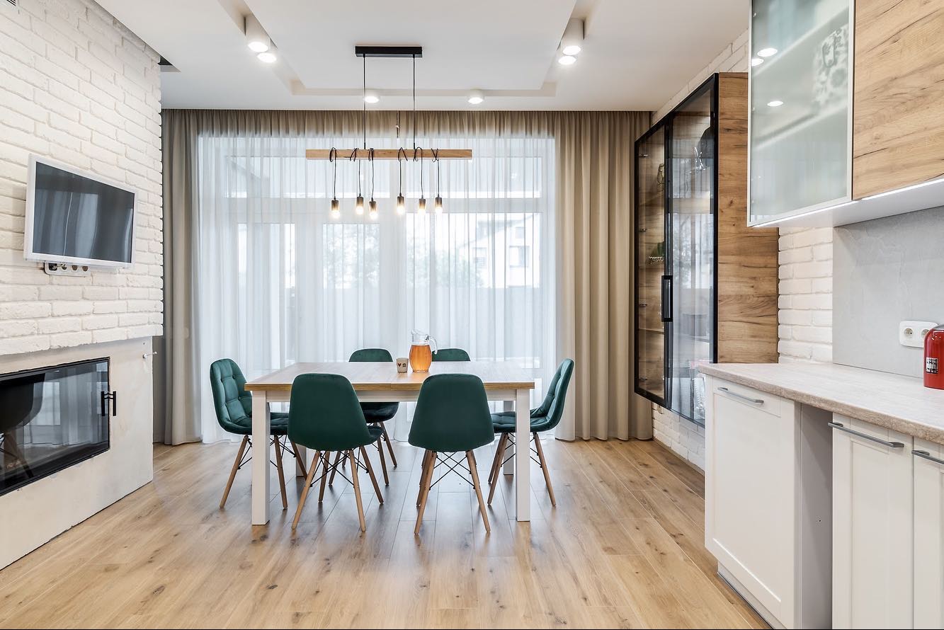 Jasne wnętrze jadalni z białymi cegłami, drewnianą podłogą i stołem z zielonymi krzesłami, oświetlone wiszącą lampą z żarówkami.