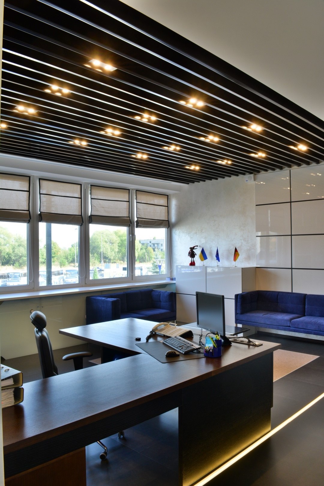 Nowoczesne biuro z ciemnym biurkiem, niebieskimi sofami i oryginalnym sufitem z podświetleniem punktowym. Na biurku komputer, telefon i przybory biurowe. W tle widoczne okna z roletami rzymskimi...