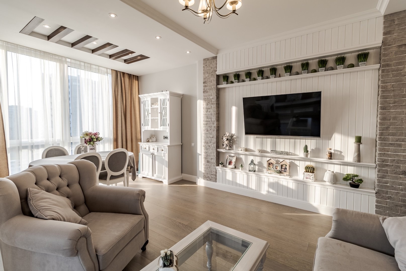 Jasny salon w stylu klasycznym z elementami rustykalnymi: beżowy fotel, biały kredens, stół z kwiatami i ścianka RTV z ceglanym akcentem i półkami dekoracyjnymi, oświetlony promieniami słonecznymi.