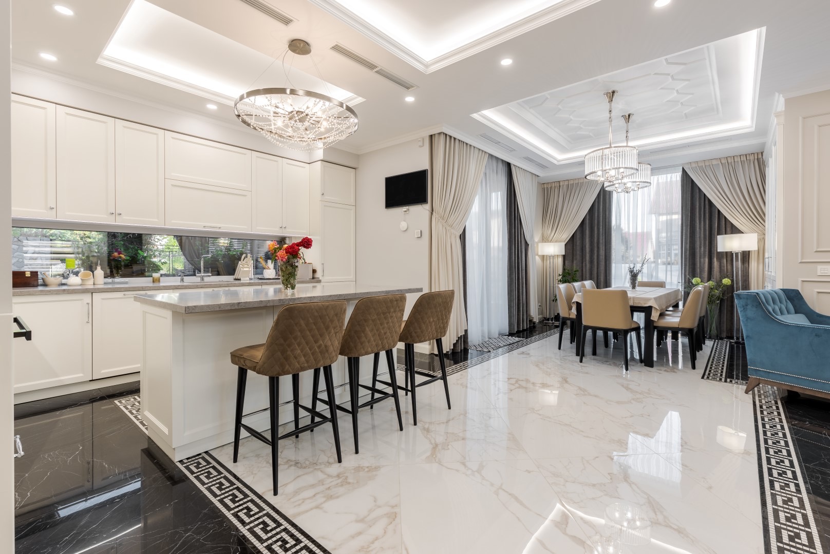 Eleganckie wnętrze: kremowa kuchnia z wyspą, brązowe krzesła barowe, marmurowa podłoga z dekoracyjnym obramowaniem, kryształowe żyrandole i widok na jadalnię z niebieskim fotelem.