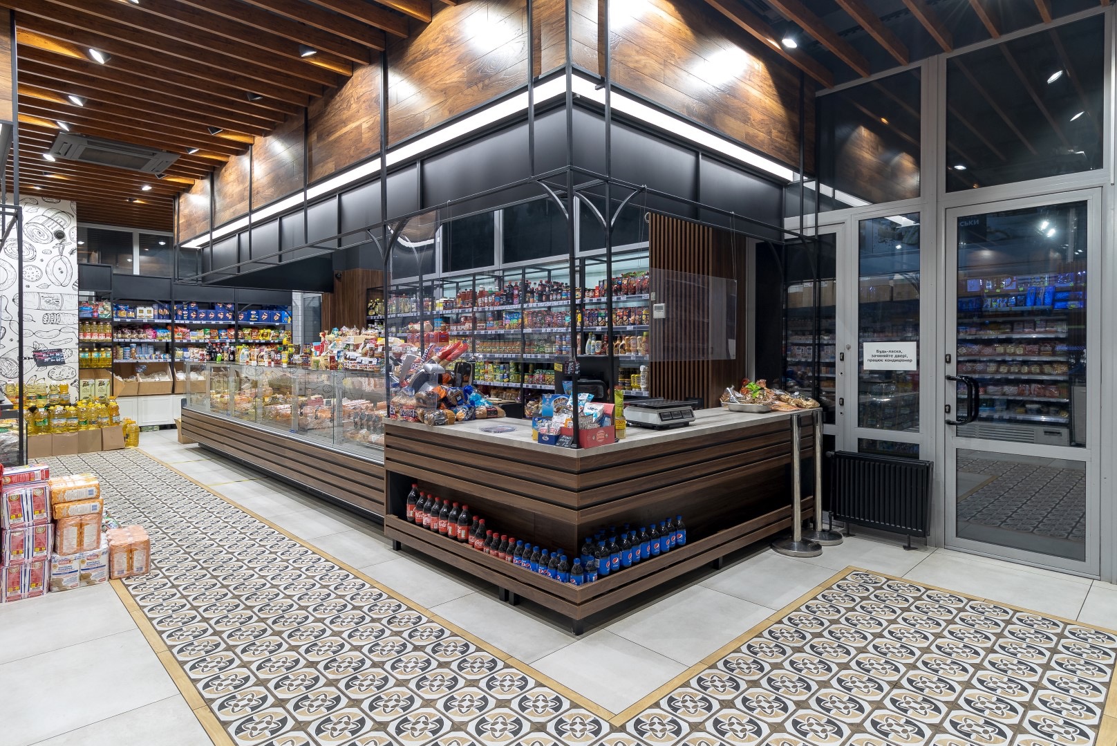 Wnętrze sklepu spożywczego z drewnianymi elementami wykończeniowymi, przeszklonymi witrynami chłodniczymi, kasą z wagą, dekoracyjną podłogą i ścianą z rysunkami produktów spożywczych.