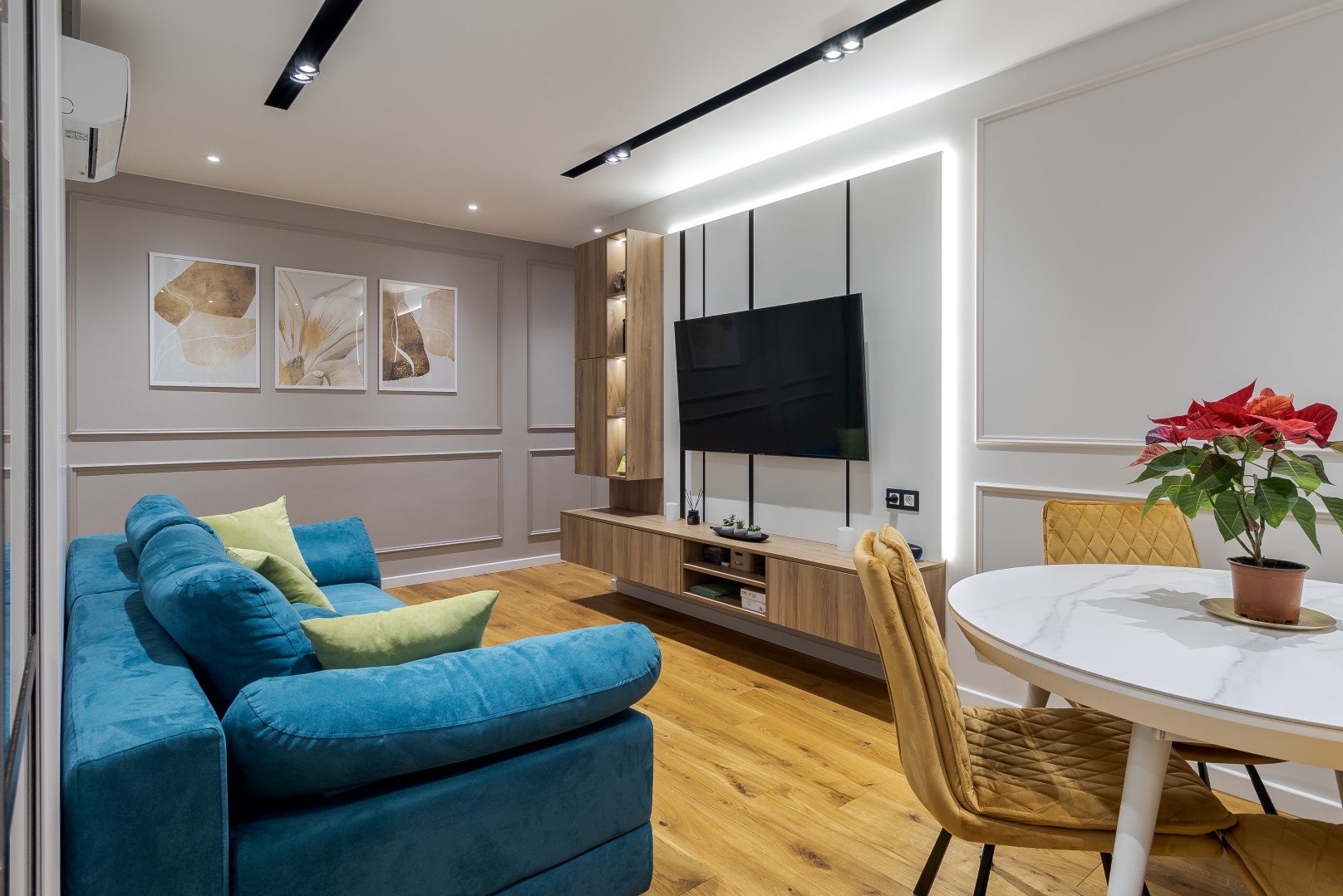 Salon z turkusową sofą i zielonymi poduszkami, drewnianą szafką RTV z półkami, telewizorem na ścianie z podświetleniem LED, okrągłym stołem z marmurowym blatem i welurowymi krzesłami w kolorze...