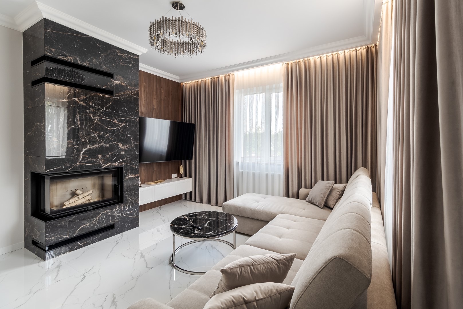 Nowoczesny salon z marmurowym kominkiem, beżową sofą narożną i okrągłym stolikiem kawowym z czarnym blatem, oświetlony dekoracyjnym żyrandolem i światłem dziennym przez okno z beżowymi zasłonami.