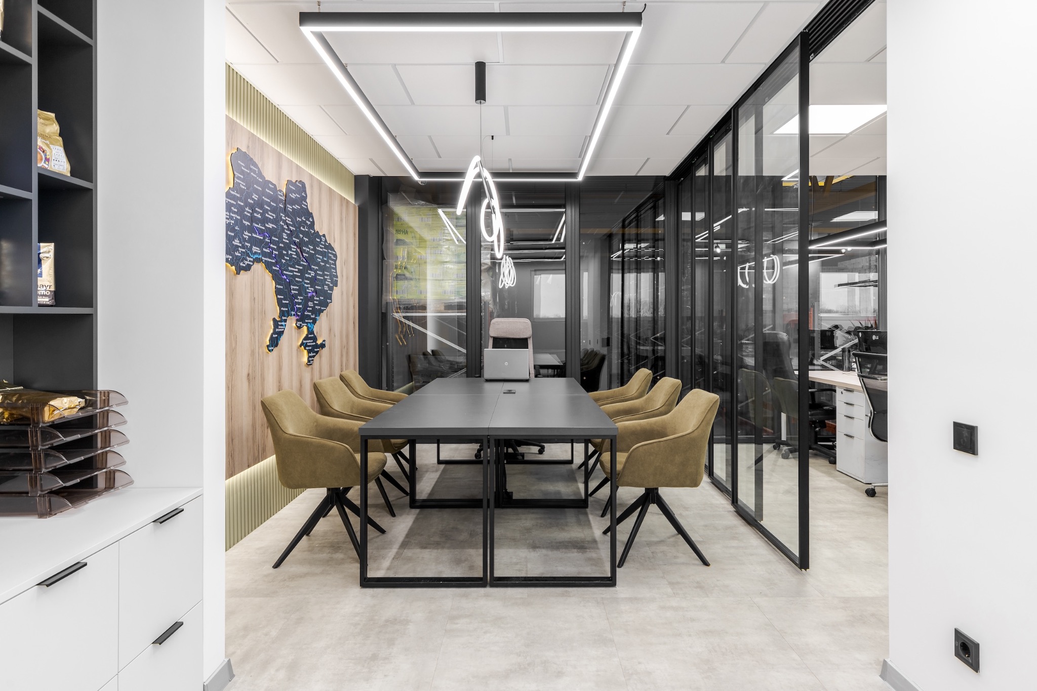 Nowoczesna sala konferencyjna z długim, szarym stołem otoczonym przez osiem krzeseł w odcieniach oliwki, laptop na stole, geometryczne oświetlenie sufitowe i mapa świata na drewnianej ścianie.