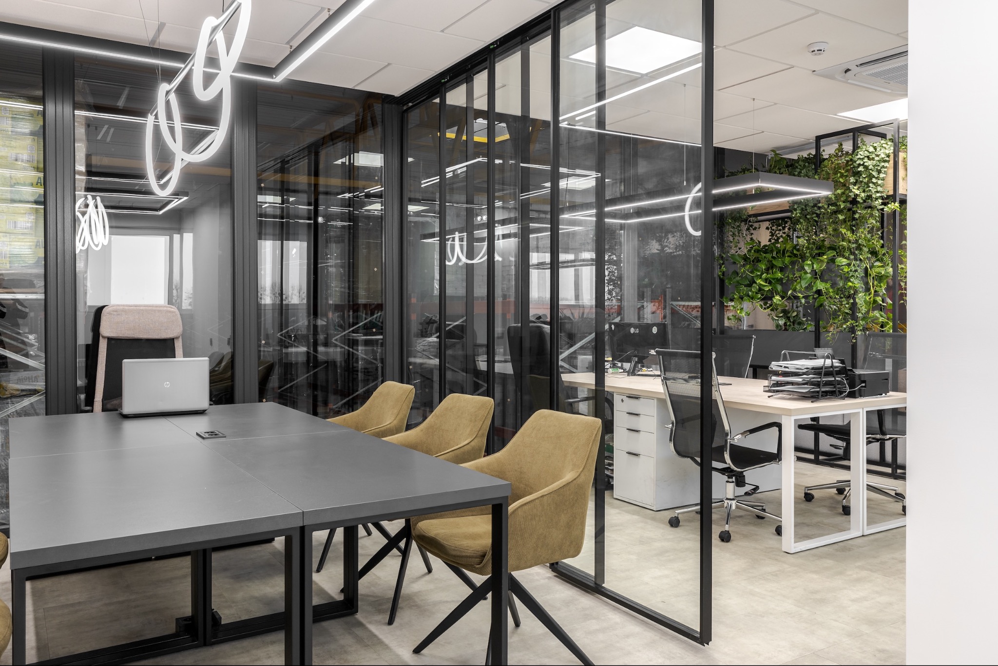 Nowoczesne biuro z przeszklonymi ścianami, szarym stołem konferencyjnym, beżowymi krzesłami, wiszącymi lampami o abstrakcyjnym kształcie i ścianą z zielonych roślin.
