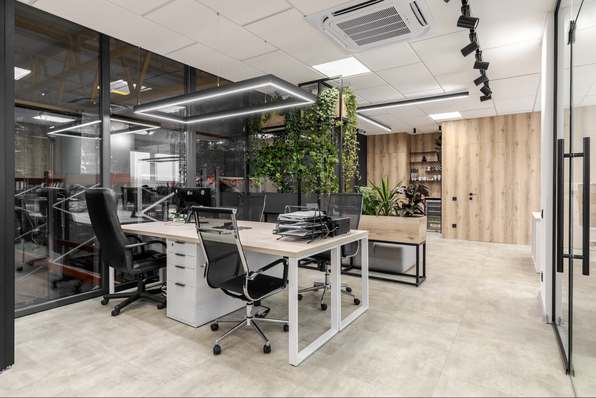 Nowoczesne biuro z biurkami, krzesłami obrotowymi i zielenią w donicach, oświetlone ledowymi lampami sufitowymi, widok z perspektywy wejścia.
