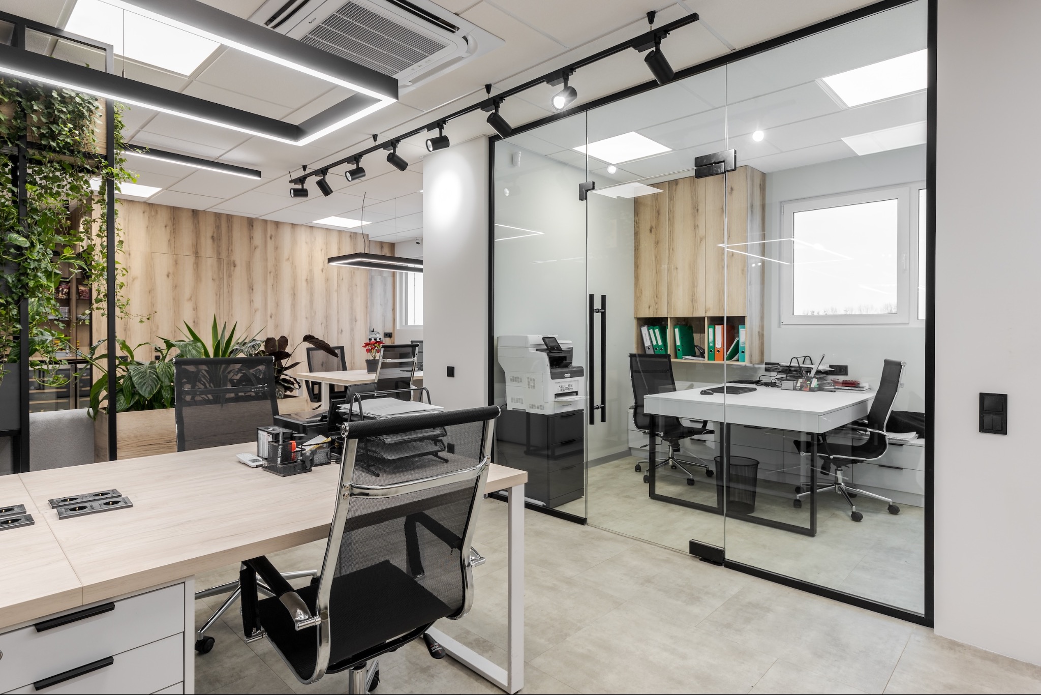 Nowoczesne biuro z przeszkloną ścianą działową, drewnianymi akcentami i zielenią, oświetlone listwami LED i reflektorami punktowymi.