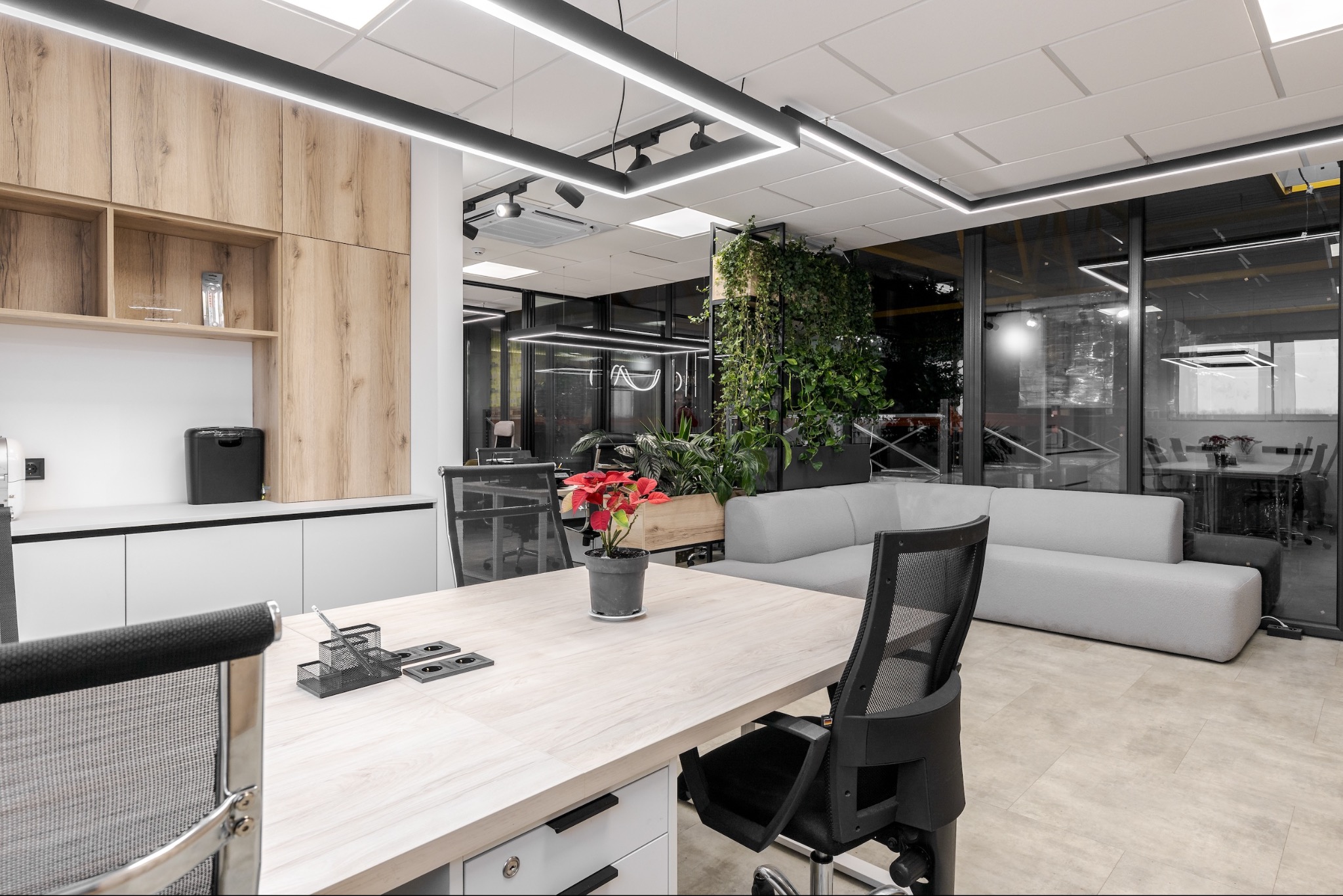 Nowoczesne biuro z jasnym biurkiem, krzesłami z siatki, roślinami doniczkowymi i sofą w strefie relaksu, oświetlone liniowymi lampami LED.