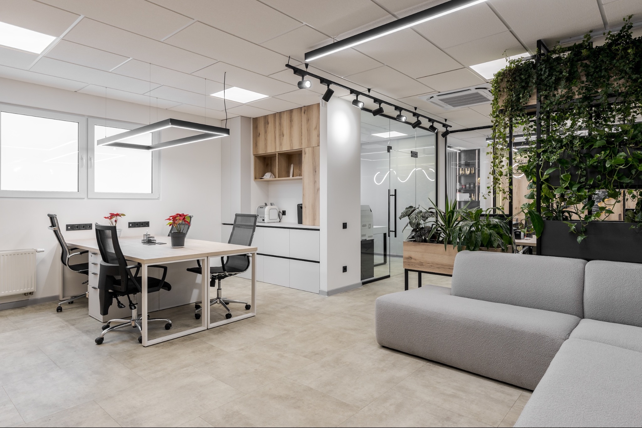 Nowoczesne biuro z minimalistycznymi biurkami, szarą sofą, roślinami w donicach, szklaną ścianą i oświetleniem LED.