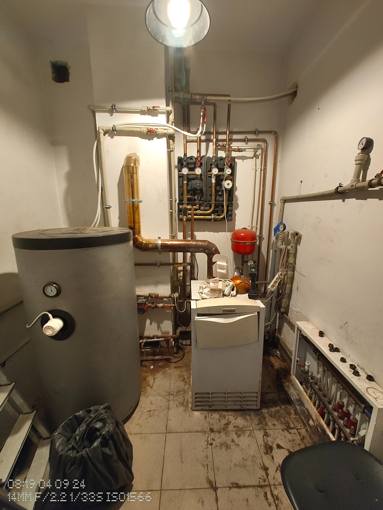 Kotłownia przed remontem: widoczny stary piec, zbiornik na wodę, rozdzielacz grzejnikowy i liczne rury. Na podłodze widoczne zabrudzenia.