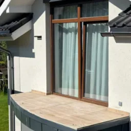 Nowoczesny balkon z drewnopodobną okładziną, czarne obramowanie, duże okno z brązowymi ramami i zasłonami, elewacja w jasnym odcieniu.