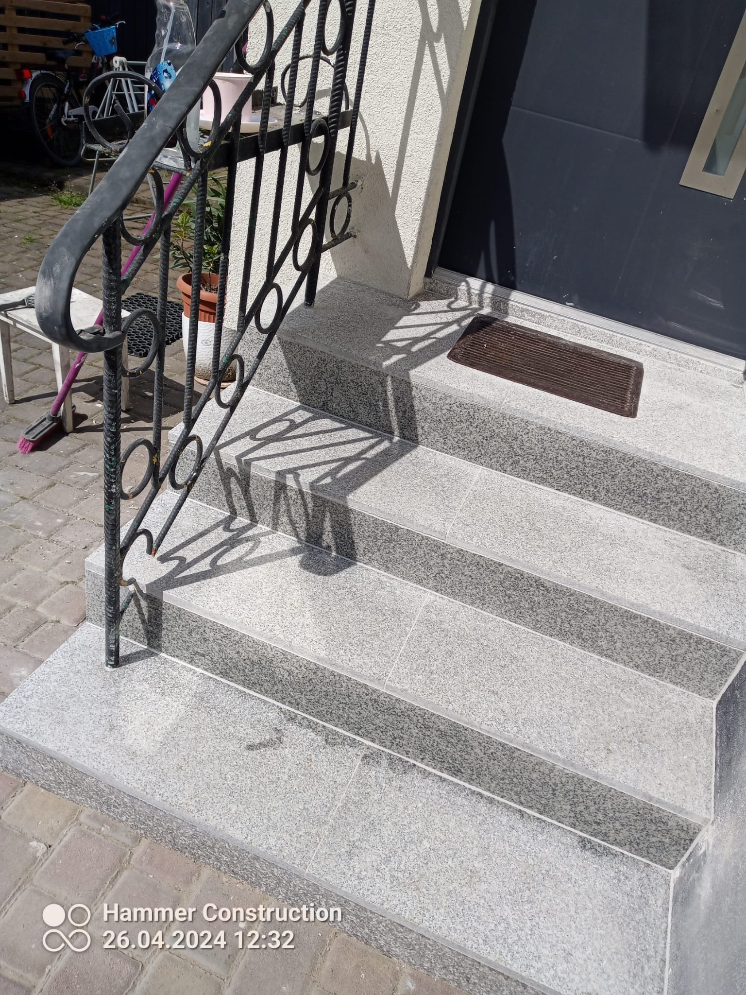 Granitowe schody wejściowe z czarną, kutą balustradą i wycieraczką przed ciemnymi drzwiami wejściowymi.