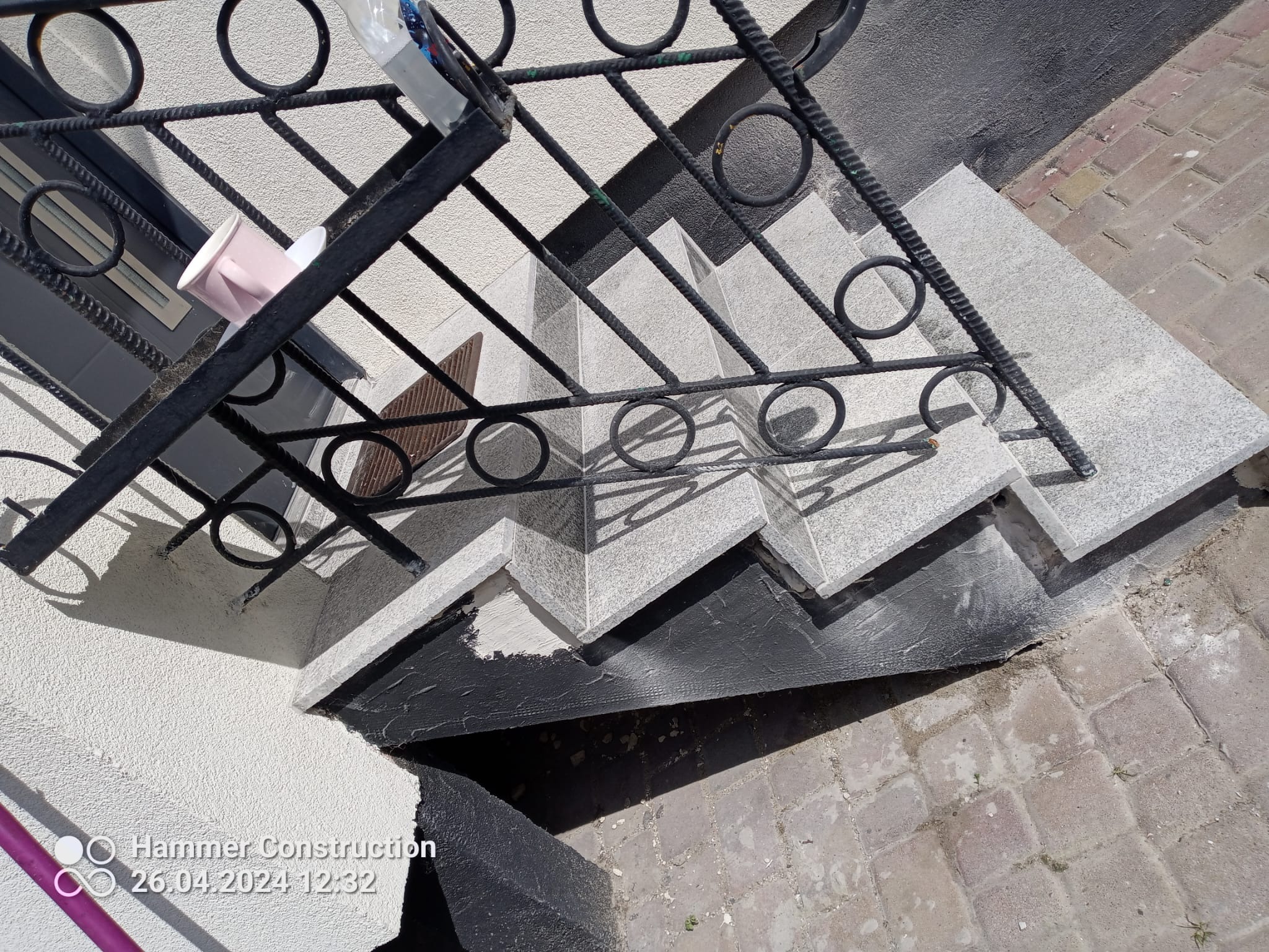 Granitowe schody zewnętrzne z czarną, metalową balustradą z okrągłymi zdobieniami, widok z góry, na schodach stoi różowy kubek i butelka wody.