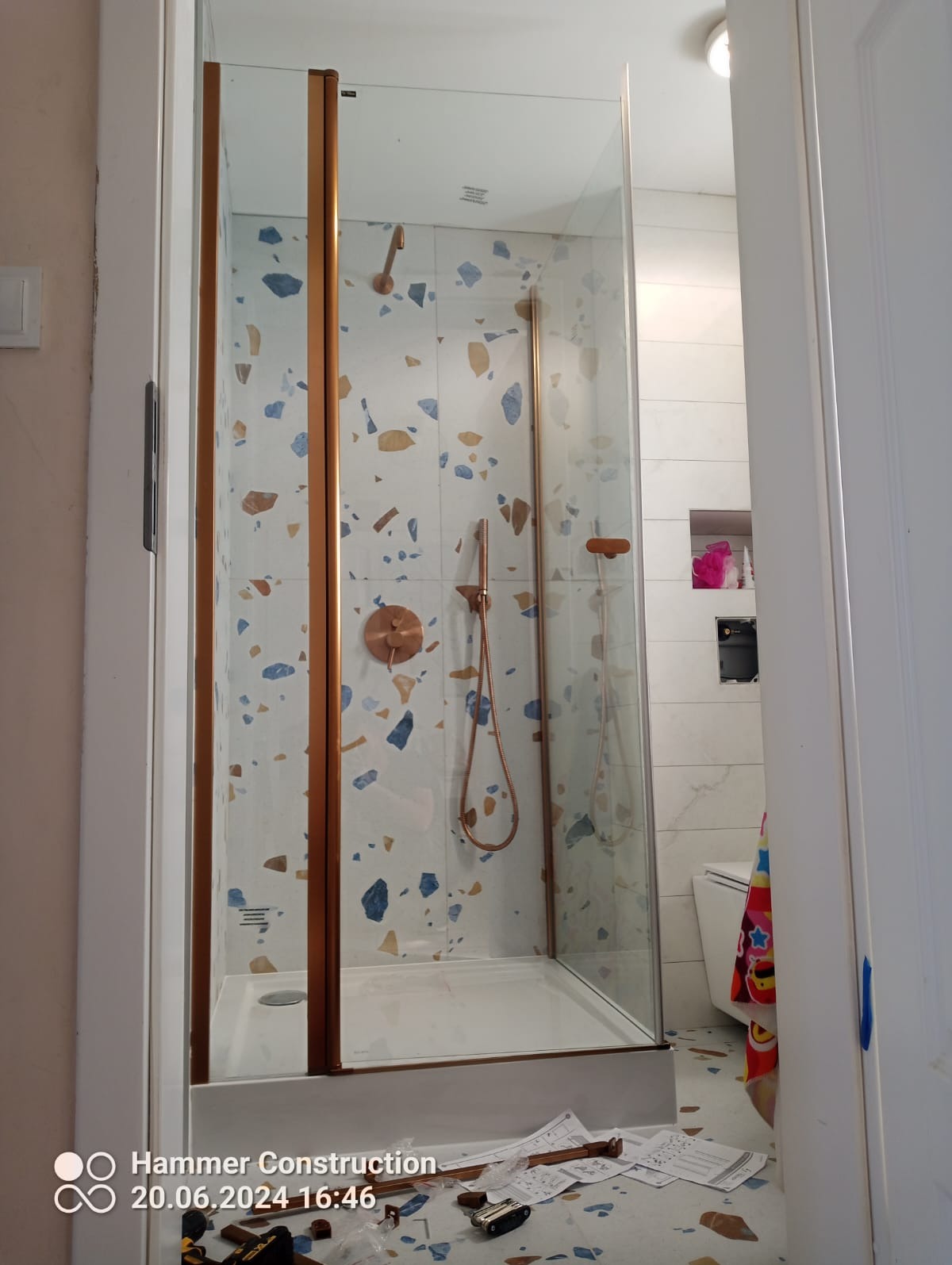 Kabina prysznicowa z elementami w kolorze miedzianym i wzorem lastryko na ścianach, widoczna zza otwartych drzwi, z narzędziami i instrukcjami na podłodze.