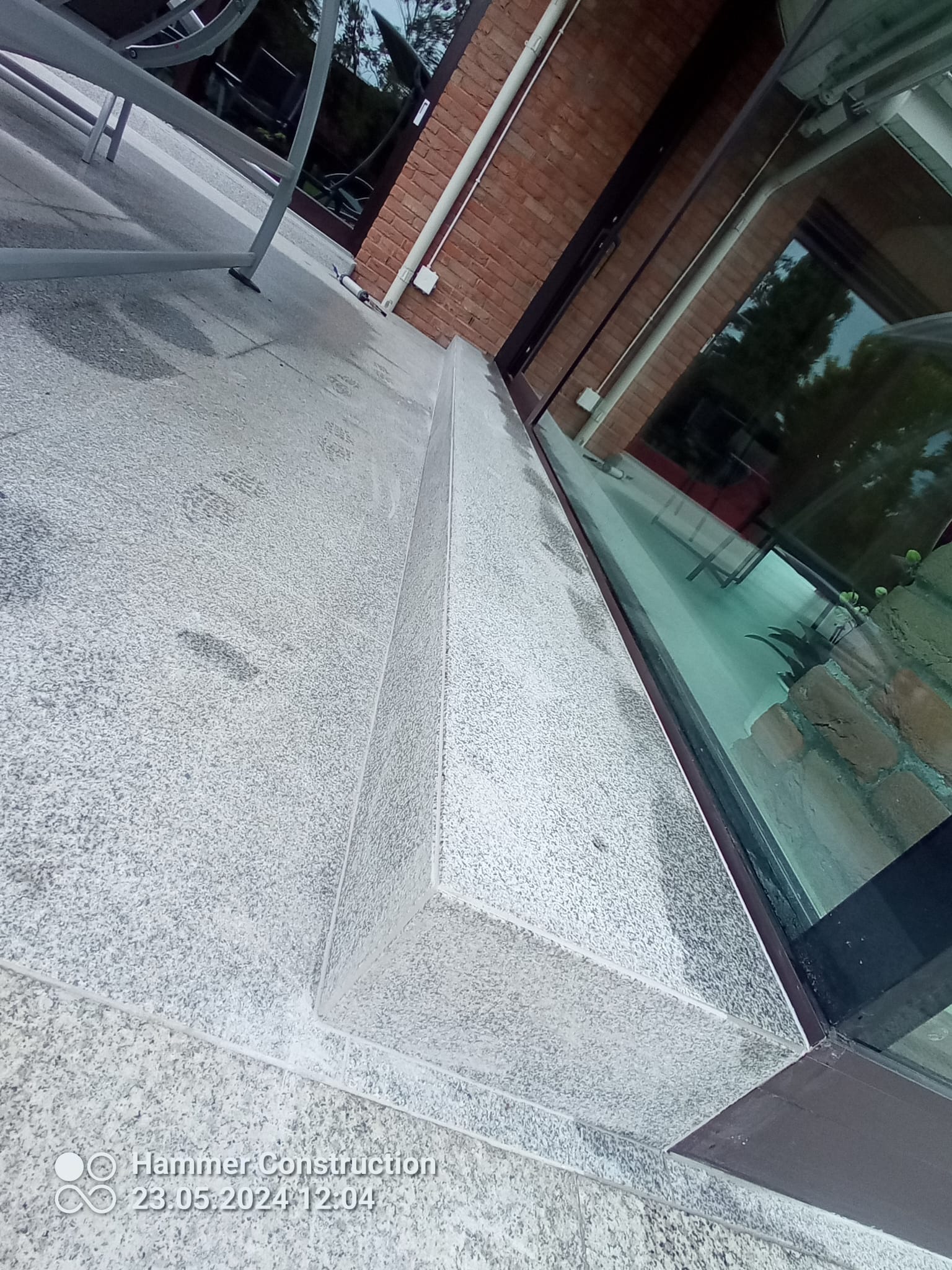 Szara, granitowa posadzka tarasu przylegająca do ceglanej ściany budynku z dużym oknem.