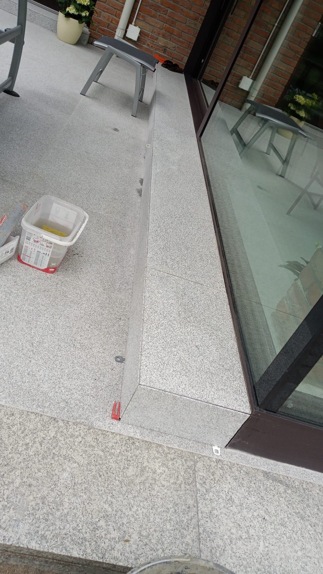 Jasnoszare płytki ułożone na tarasie przy oknie, widoczne narzędzia glazurnicze: wiadro z zaprawą, poziomica i krzyżyki dystansowe.