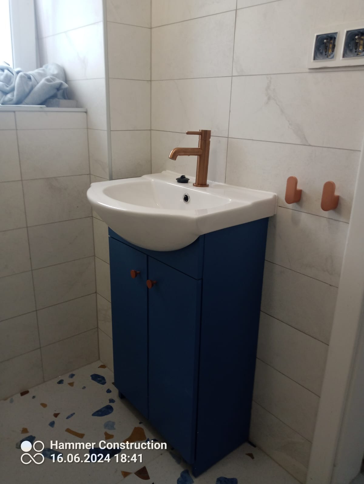 Nowoczesna łazienka z białą umywalką nablatową na granatowej szafce, miedziana bateria, płytki imitujące marmur na ścianach oraz podłoga lastryko.