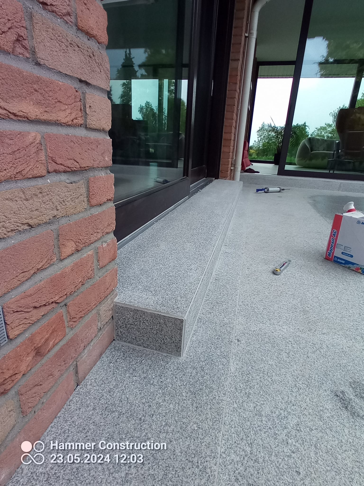 Wykończone schody zewnętrzne z jasnoszarego kamienia, przylegające do ceglanej ściany budynku z dużymi oknami; widoczne narzędzia i opakowanie po materiałach budowlanych.