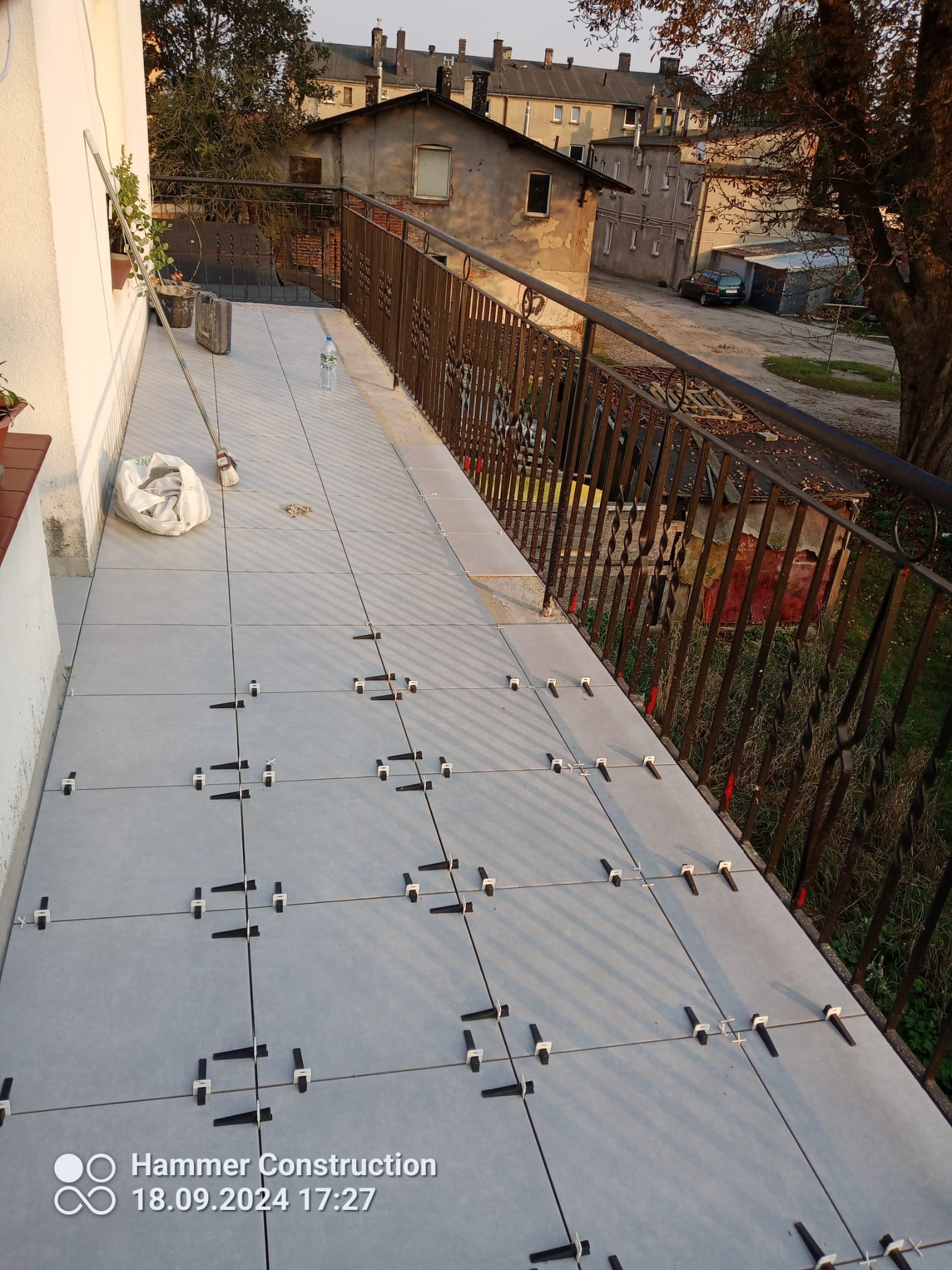 Jasnoszare płytki ułożone na balkonie z użyciem krzyżyków dystansowych, widoczna metalowa balustrada, w tle budynki mieszkalne i drzewo.