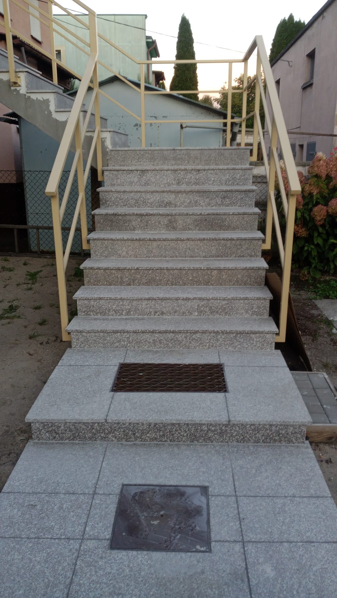 Granitowe schody zewnętrzne z żółtymi metalowymi barierkami w Starogardzie Gdańskim, prowadzące do budynku, z widocznymi kratkami odpływowymi.