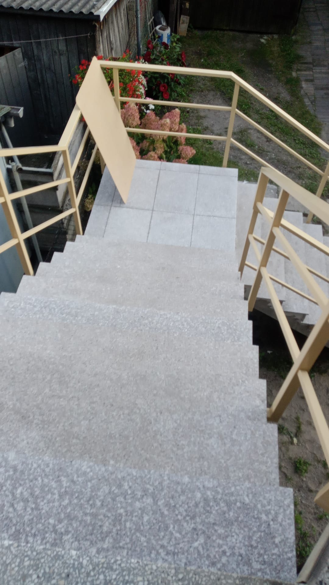 Zewnętrzne schody z szarego lastryko, z beżową metalową balustradą, widok z góry na dół, fragment ogrodu z kwiatami w tle.