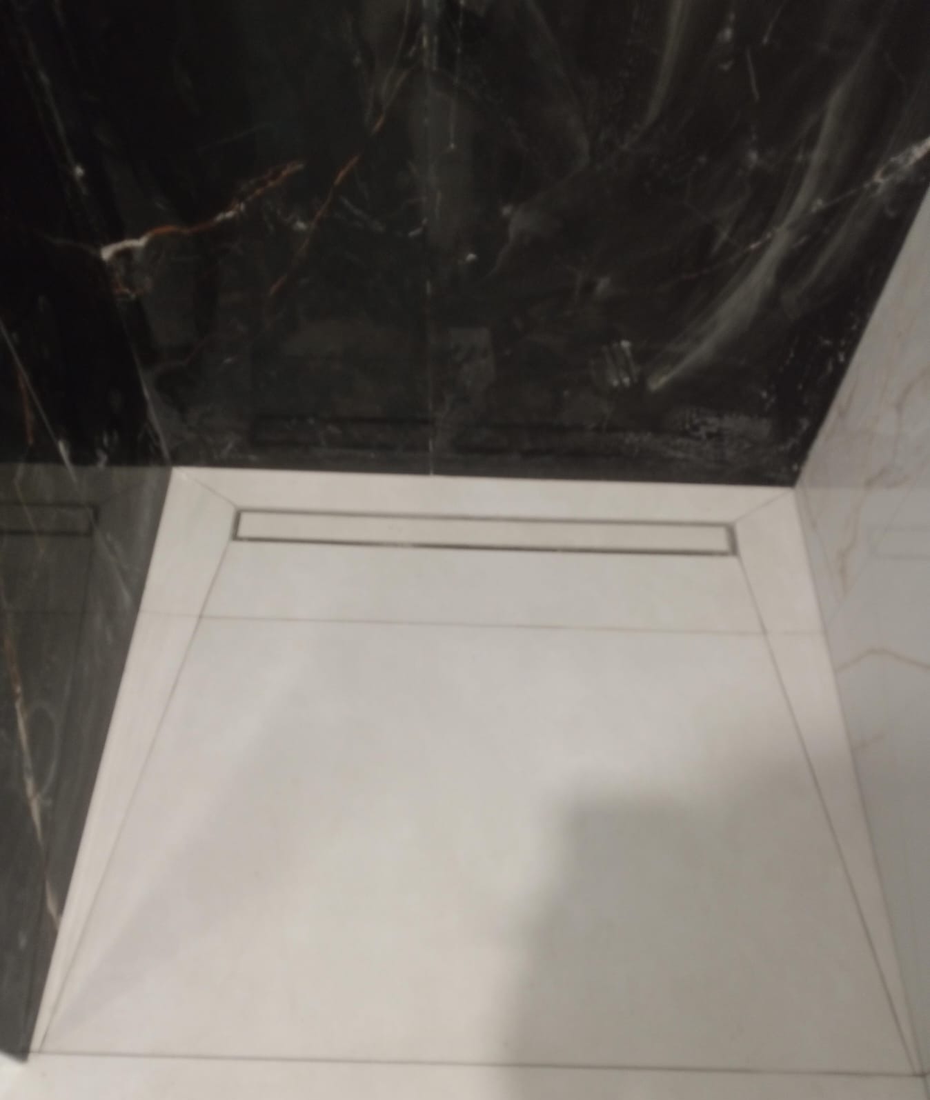 Nowoczesny odpływ liniowy w kabinie prysznicowej wyłożonej białymi płytkami podłogowymi i czarnymi płytkami imitującymi marmur na ścianach.