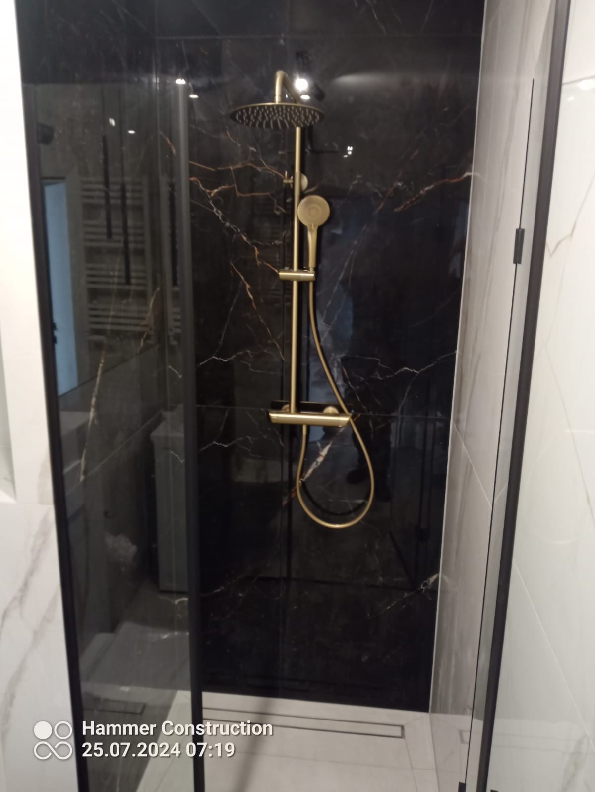 Kabina prysznicowa z czarnymi płytkami imitującymi marmur i złotą armaturą, widoczny odpływ liniowy w podłodze.